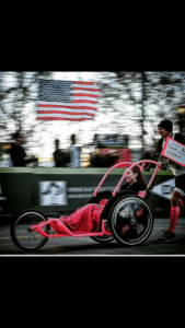 Man pushing a girl in a pink racing wheelchair with the text "Let's Roll, Baby-girl."